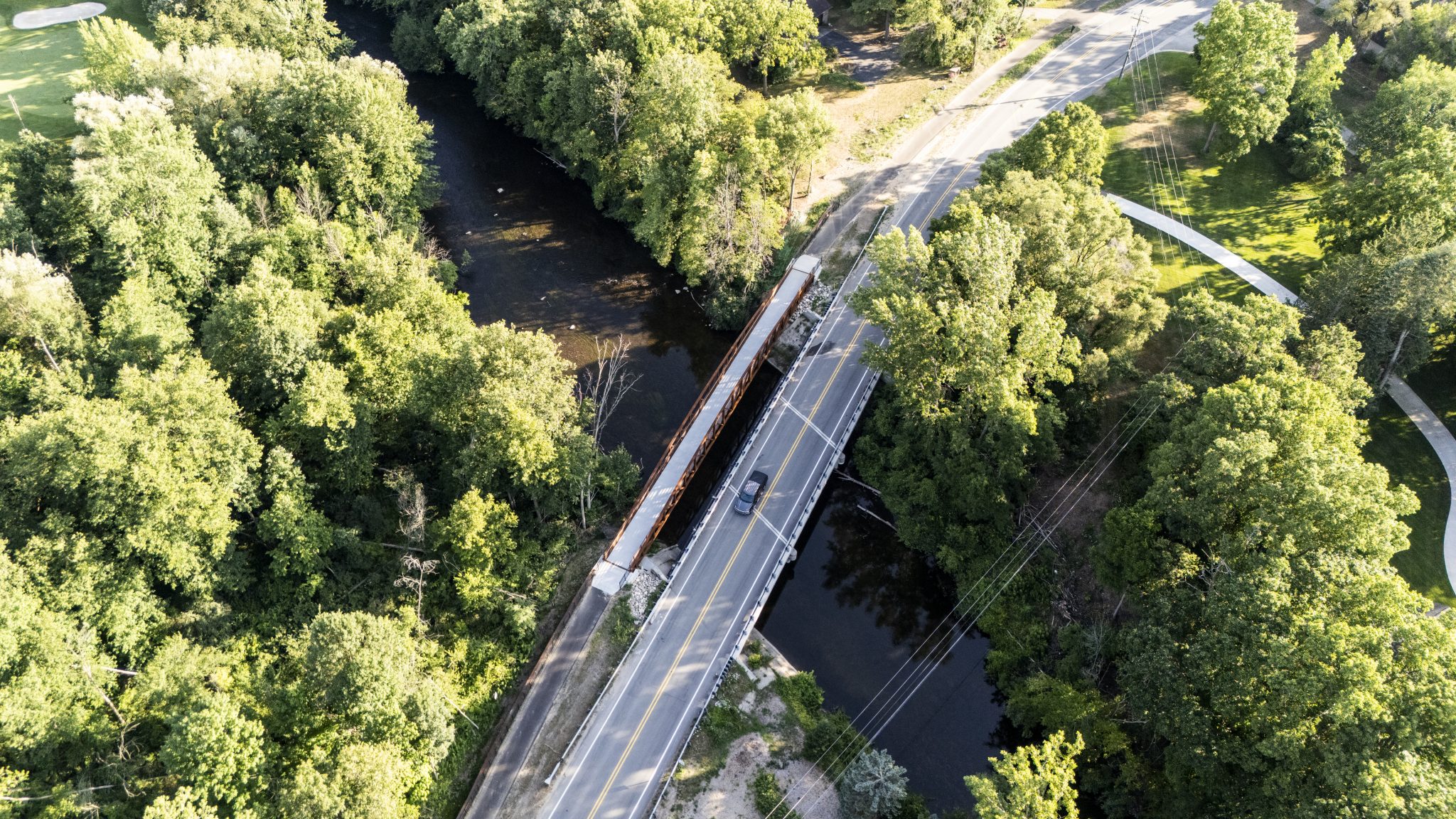 Drone view of a pedestrian bridge alongside a motor road over a river surrounded by trees