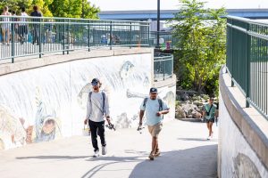 A pair of anglers walking up a busy riverwalk next to a public mural with background greenery