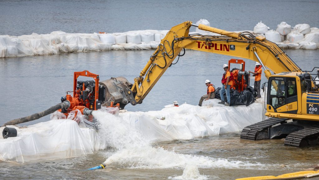 A yellow truck dewatering a section in a river surrounded by white sandbags and construction works